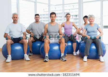 Portrait of fitness class with dumbbells sitting on exercise balls in a bright gym