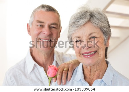 Senior man offering a rose to his partner smiling at camera at home in living room
