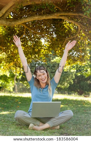 Full length portrait of a cheerful young woman with laptop raising hands at the park