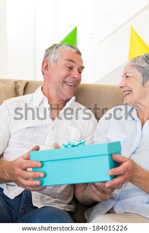 Senior couple sitting on couch wearing party hats holding a gift at home in living room