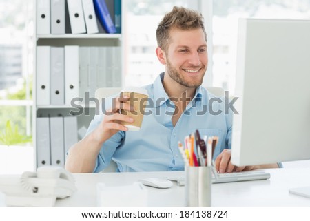 Smiling man working at his desk drinking a take away coffee in creative office