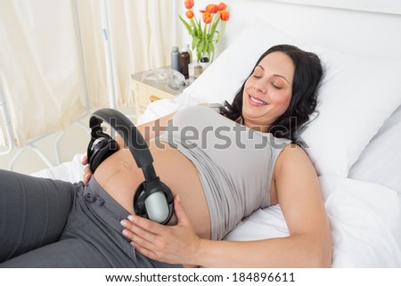 Smiling expectant woman putting headphones on her belly while lying in bed at hospital