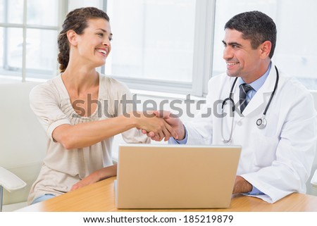 Male doctor and patient shaking hands by laptop at desk in medical office