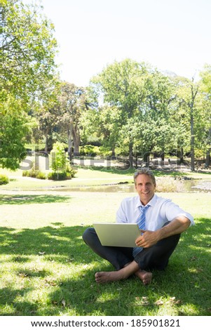 Full length portrait of smiling businessman with laptop sitting on grass in park