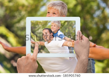 Hand holding tablet pc showing father giving son a piggy back in park