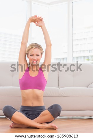 Happy blonde sitting in lotus pose stretching arms up at home in the living room