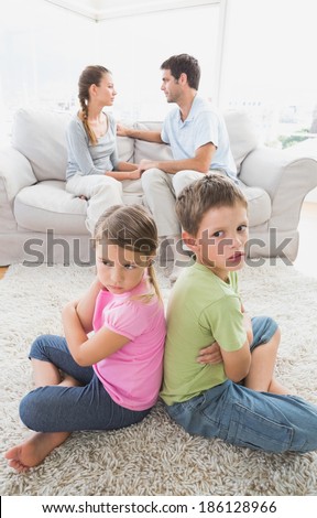 Pouting siblings sitting back to back while parents are arguing at home in the living room