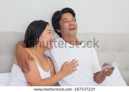 Smiling asian couple lying on bed watching tv at home in bedroom