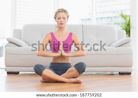 Calm blonde sitting in lotus pose with hands together at home in the living room
