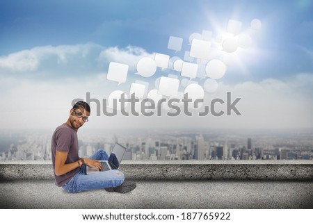 Man wearing glasses sitting on floor using laptop and looking at camera against balcony overlooking city