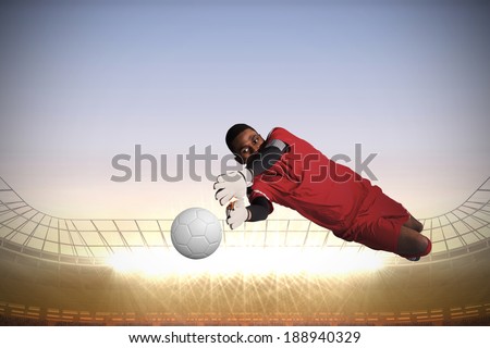 Goalkeeper in red making a save against large football stadium with spotlights