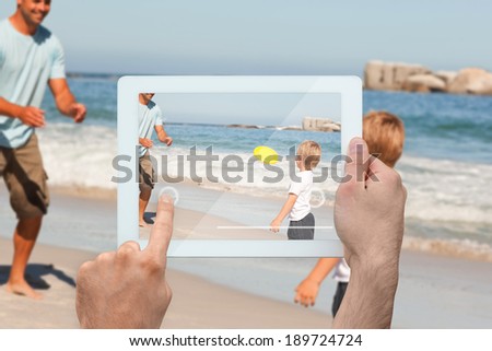 Hand holding tablet pc showing father playing frisbee with son at beach