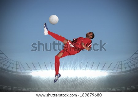 Football player in red kicking against large football stadium with spotlights under grey sky