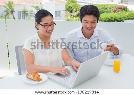 Smiling couple having breakfast together using laptop outside on a balcony
