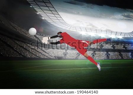 Goalkeeper in red making a save in a large football stadium with lights