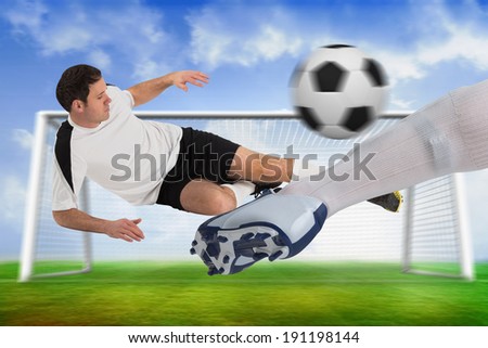 Composite image of football players tackling for the ball against football pitch and goal under blue sky