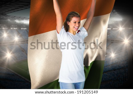 Pretty football fan in white cheering holding ivory coast flag against large football stadium with fans in blue