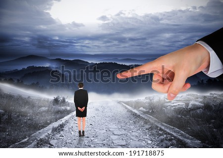 Giant hand pointing at businesswoman standing with hands behind back against stony path leading to large misty mountains