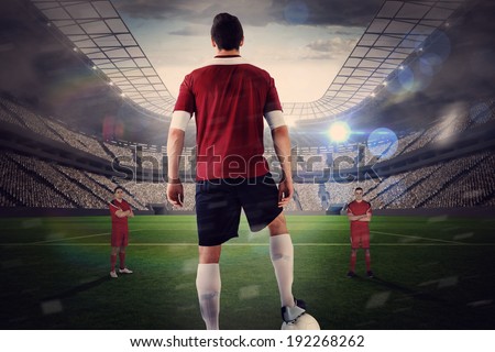 Football player in red with ball facing opposition against large football stadium with lights