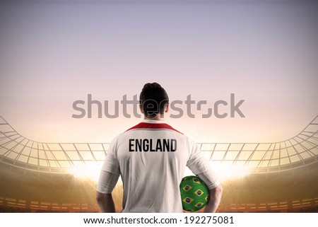 England football player holding ball against large football stadium under blue sky