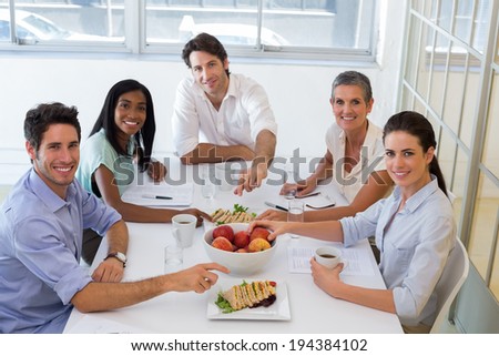 Business people smiling at camera eating sandwiches and fruit for lunch in the office