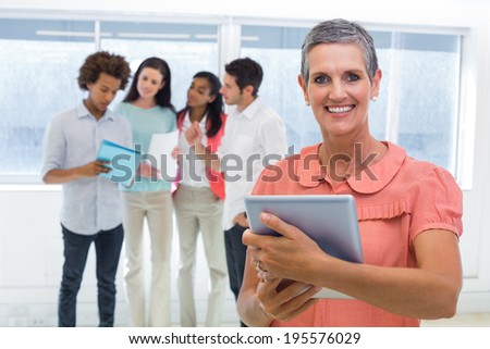 Businesswoman smiles at camera while looking at tablet pc with coworkers behind her in the office
