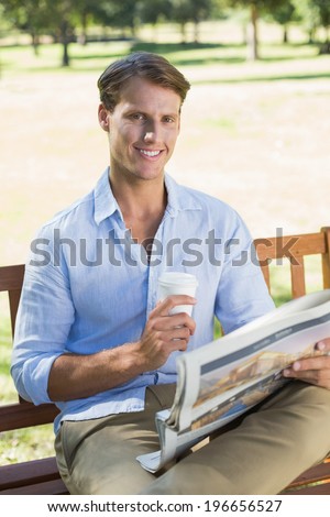 Smiling man sitting on park bench drinking coffee and reading paper on a sunny day