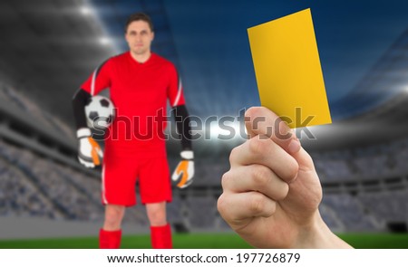 Hand holding up yellow card against football stadium with goalie