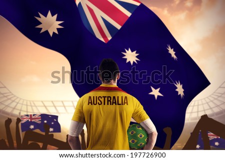 Australia football player holding ball against large football stadium under cloudy blue sky