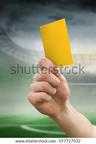 Hand holding up yellow card against football pitch in large stadium