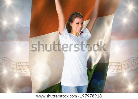 Pretty football fan in white cheering holding ivory coast flag against large football stadium under cloudy blue sky