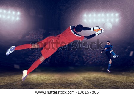 Goalkeeper in red making a save against large football stadium under blue sky
