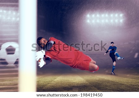 Goalkeeper in red making a save against large football stadium under blue sky