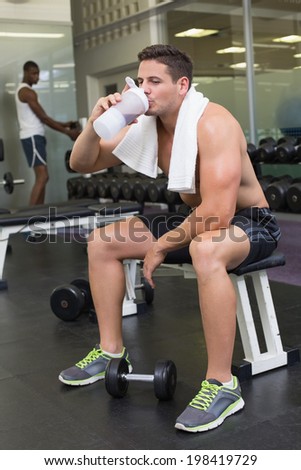 Shirtless bodybuilder holding protein drink sitting on bench at the gym