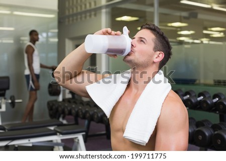 Shirtless bodybuilder drinking protein drink sitting on bench at the gym