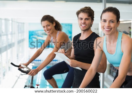 Fit women in a spin class with trainer taking notes and smiling at camera at the gym