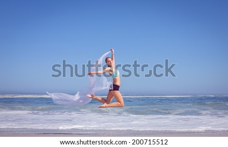 Fit woman jumping gracefully on the beach with scarf on a sunny day