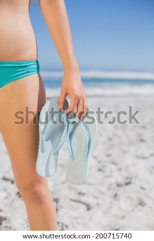 Mid section of fit woman in bikini on beach holding flip flops on a sunny day