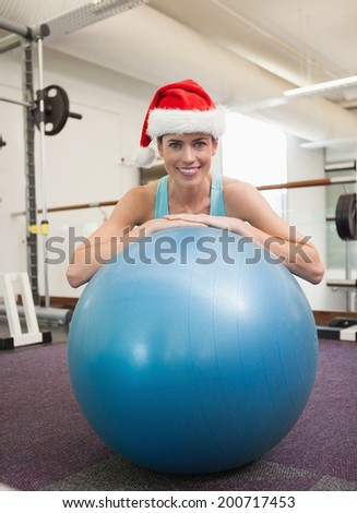 Fit smiling brunette in santa hat leaning on exercise ball at the gym