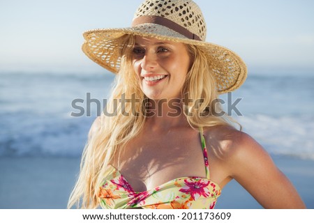 Gorgeous blonde in straw hat and bikini smiling on beach on a sunny day
