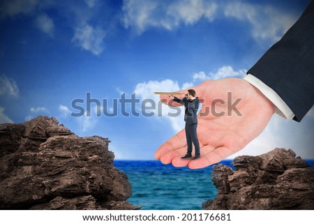 Businessman looking through telescope in large hand against large rocks overlooking sea and sky