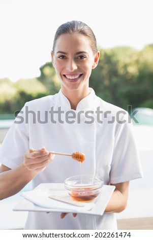 Smiling beauty therapist looking at camera holding plate with honey outside at the spa