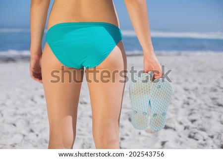 Rear view of fit woman in bikini on beach holding flip flops on a sunny day
