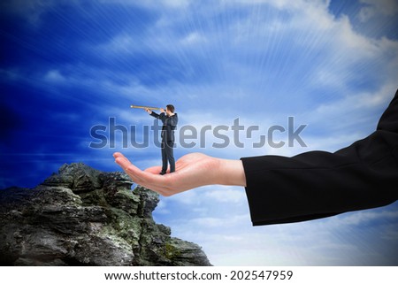 Businessman looking through telescope in hand against large rock overlooking bright blue sky