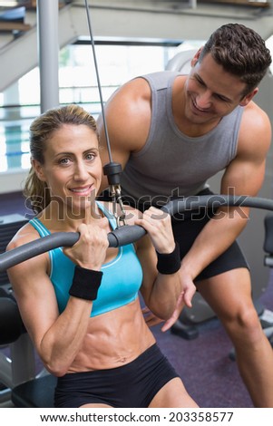 Female bodybuilder using weight machine for arms with encouraging trainer at the gym