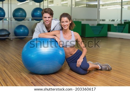 Fit woman leaning on exercise ball with trainer smiling at camera at the gym