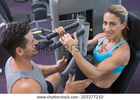 Personal trainer coaching smiling female bodybuilder using weight machine at the gym