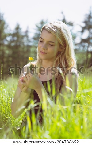 Pretty blonde in sundress sitting on grass holding yellow flower on a sunny day in the countryside