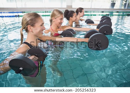 Smiling female fitness class doing aqua aerobics with foam dumbbells in swimming pool at the leisure centre
