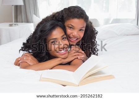 Mother and daughter reading book together on bed at home in bedroom